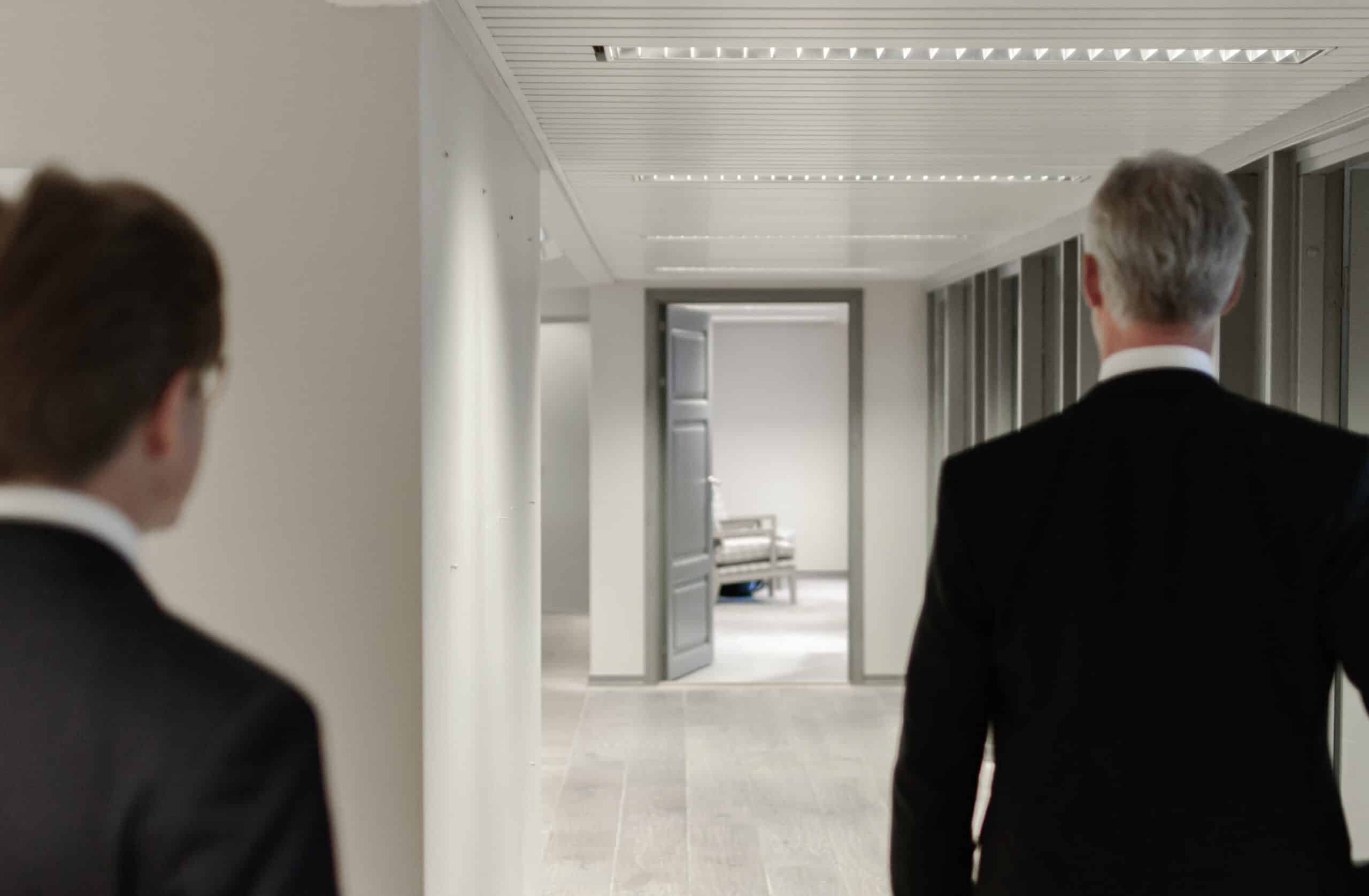 Two men in suits walk down a bright office hallway, viewed from behind.
