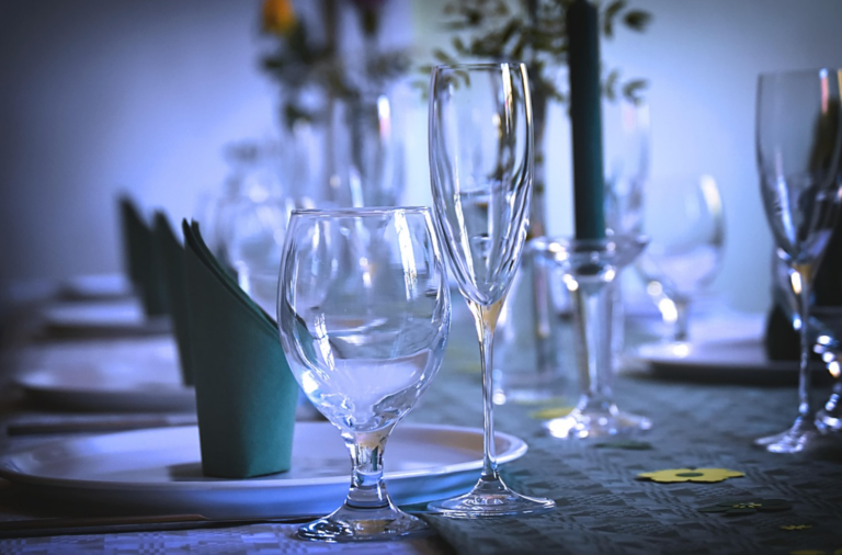 Close-up of a formal dining table with empty wine glasses, a teal napkin, and scattered confetti on the tablecloth.