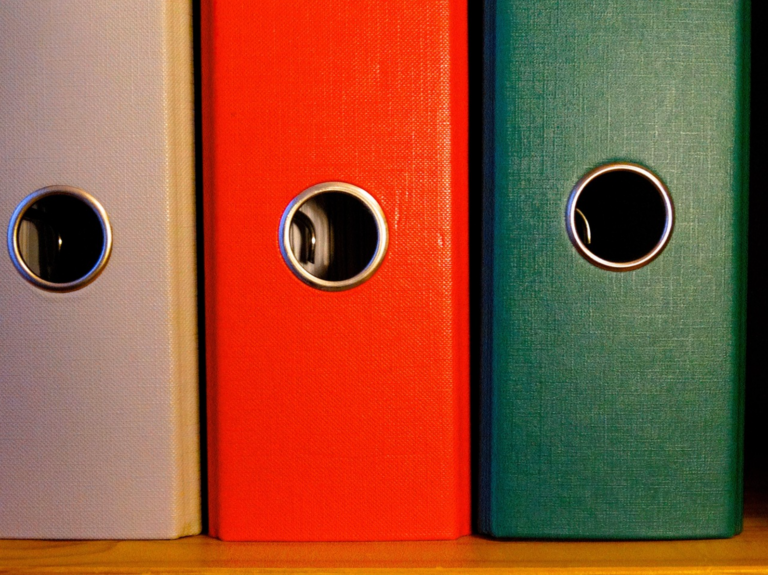 Three vertical office binders in beige, red, and teal on a wooden shelf, viewed from the side.