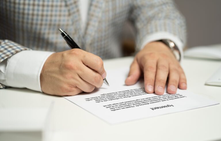 Person signs a document with a black pen at a white desk, close-up of hands on paper.